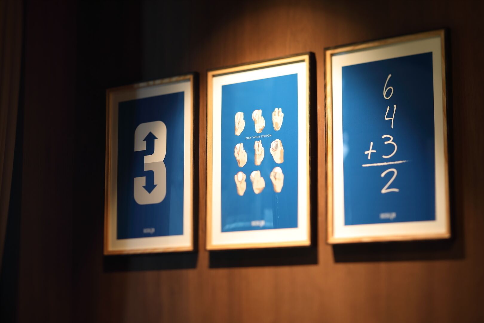 Three framed items on a wall: a large number '3', a collection of objects with 'PICK YOUR POISON.', and a mathematical expression '6 4 + 3/2'.