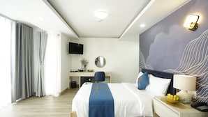 A hotel room with a large bed, a desk, a chair, a TV, a lamp, and a fruit bowl.