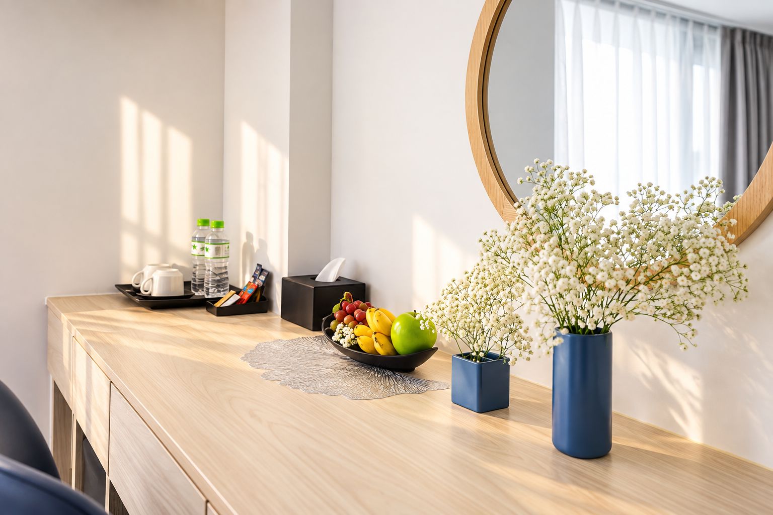 Habitación de hotel con un escritorio de madera, un espejo, un frutero, una caja de pañuelos y un jarrón con flores.