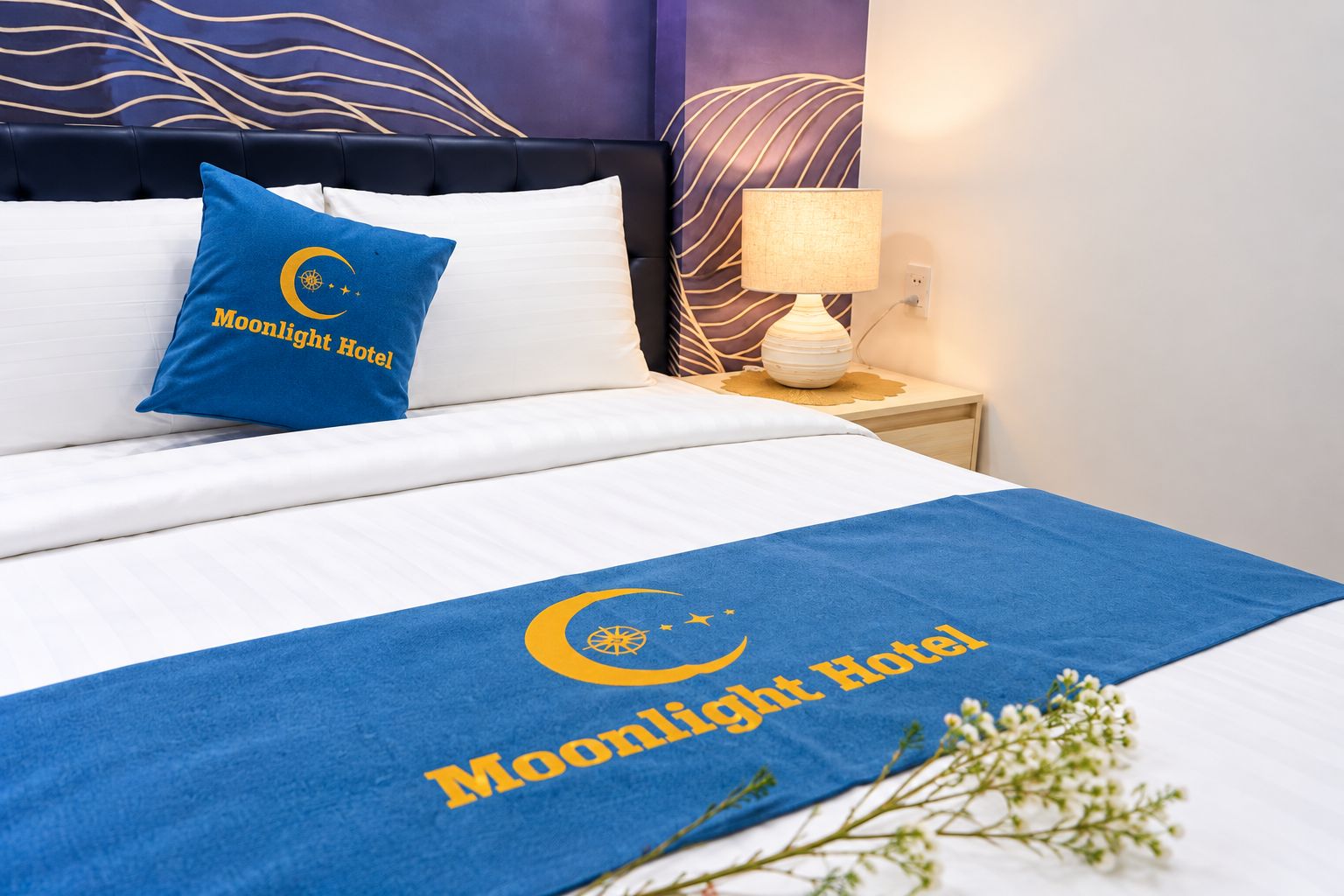 A hotel room with a bed, pillows, a nightstand, a lamp, and a decorative plant.