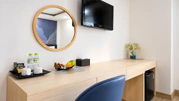 Habitación de hotel con un escritorio de madera, un televisor montado en la pared, un espejo, una pequeña planta en maceta y una bandeja con artículos de aseo y frutero.