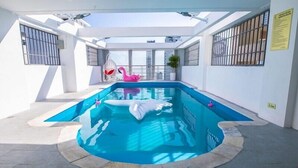 Una piscina interior con un flotador con forma de tiburón blanco, un flotador de flamenco rosa y un techo de vidrio transparente.
