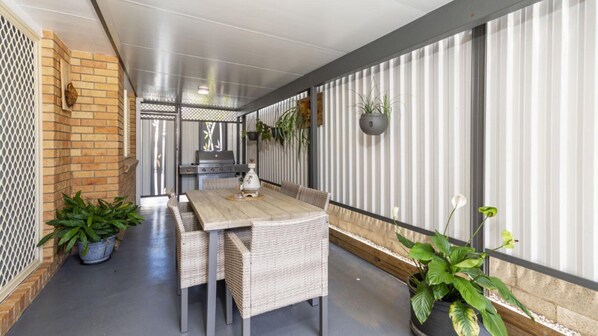 A covered outdoor dining area with a long table and wicker chairs, surrounded by potted plants and a grill.