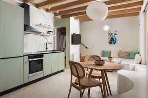 A modern kitchen with a built-in oven, a dining area with a round table and chairs, and a living space with a sofa and cushions.