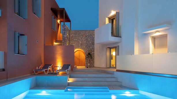 Un área de piscina exterior moderna con escalones que conducen a un muro de piedra, sillas de descanso y una entrada bien iluminada.