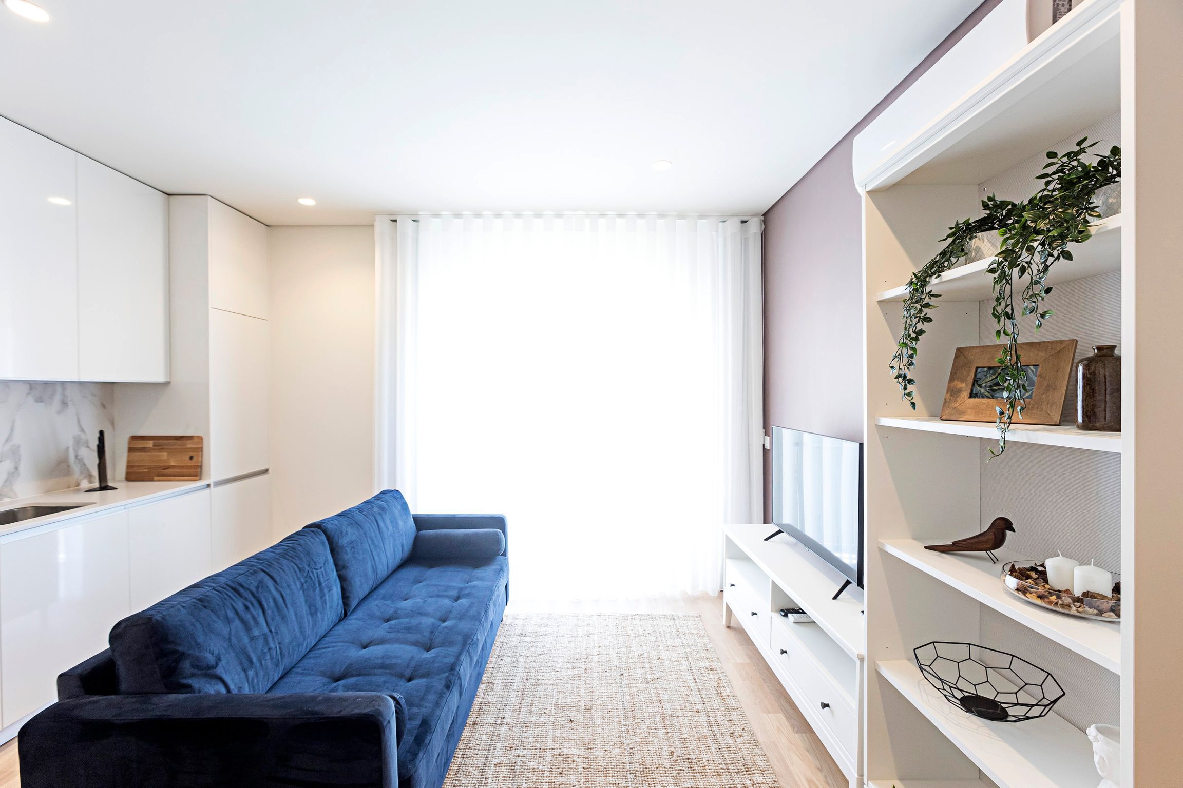 Ein modernes Wohnzimmer mit einem blauen Sofa, weißen Schränken, einem Fernseher und einer Pflanze auf Regalen.