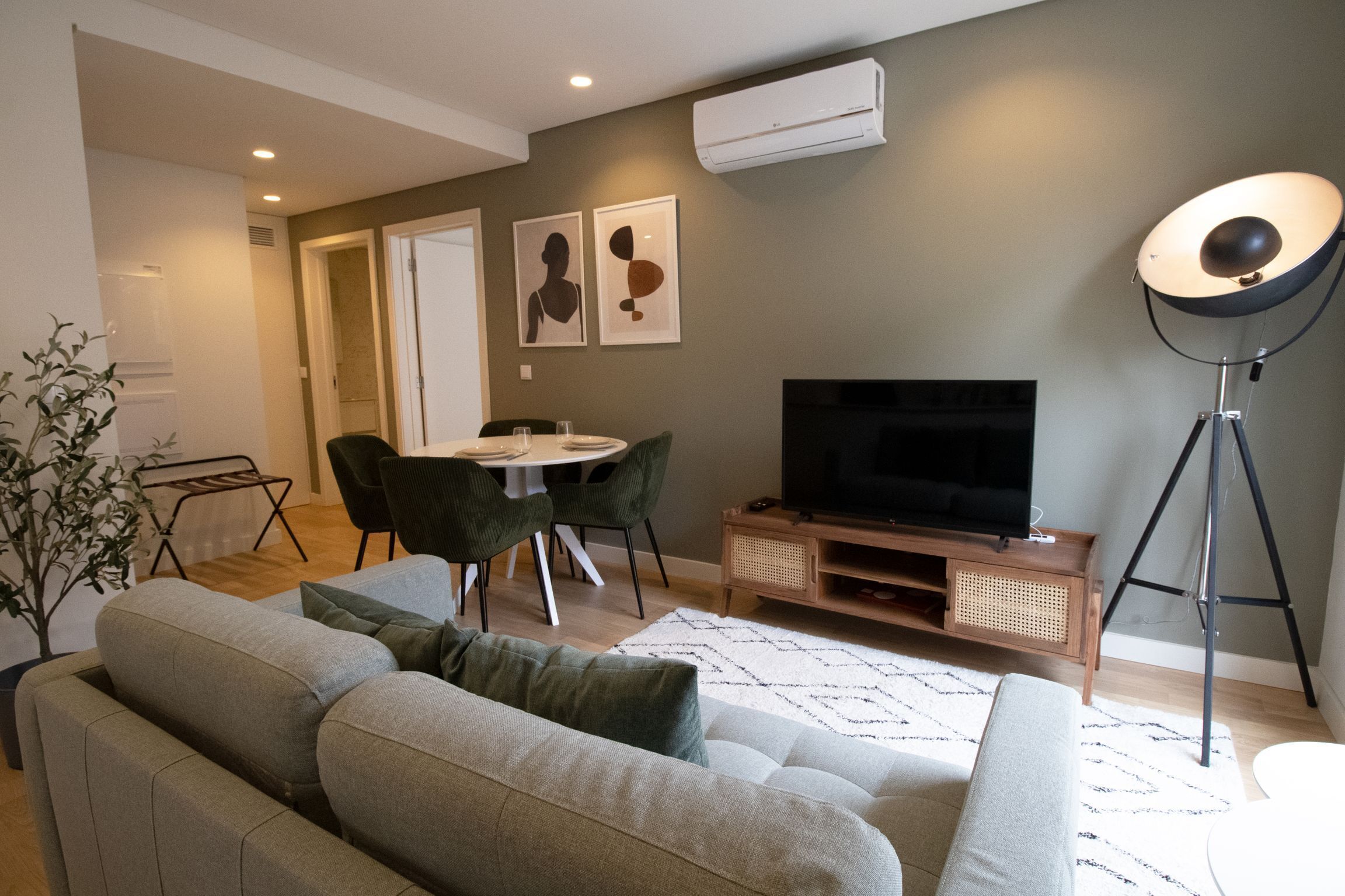 Ett modernt vardagsrum med en grå soffa, ett matbord med stolar, en platt-TV och en golvlampa.