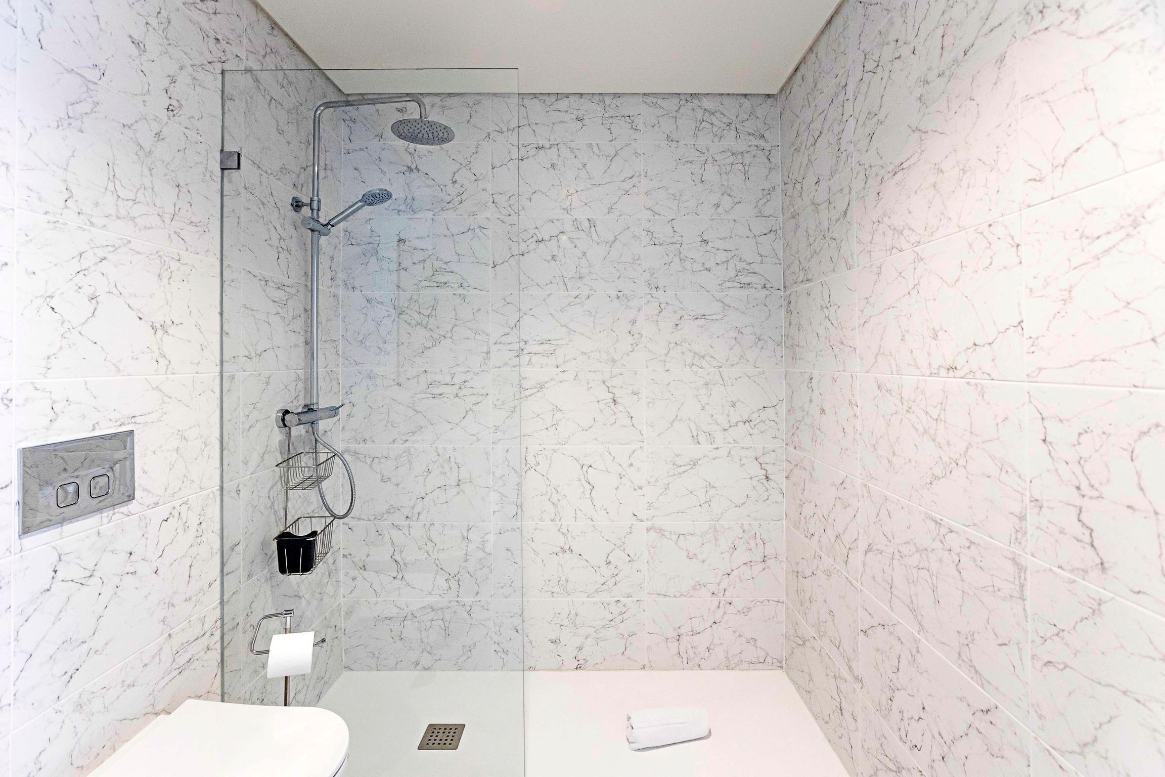 A bathroom with a glass-enclosed shower, marble walls, a white sink, and a towel on the floor.
