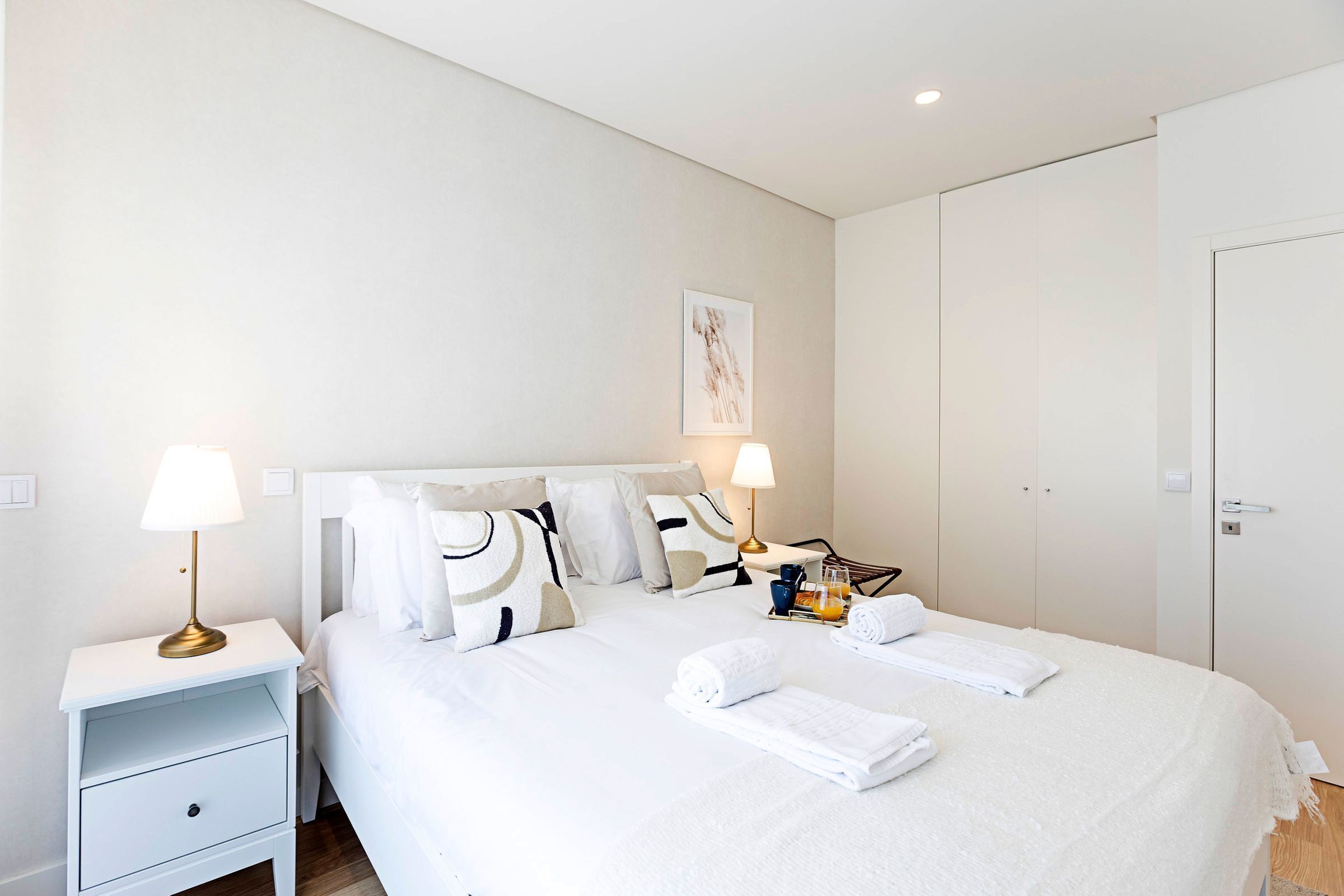 Habitación de hotel ordenada con una cama, dos mesitas de noche, una lámpara, una toalla y una pequeña mesita con una botella y vasos.