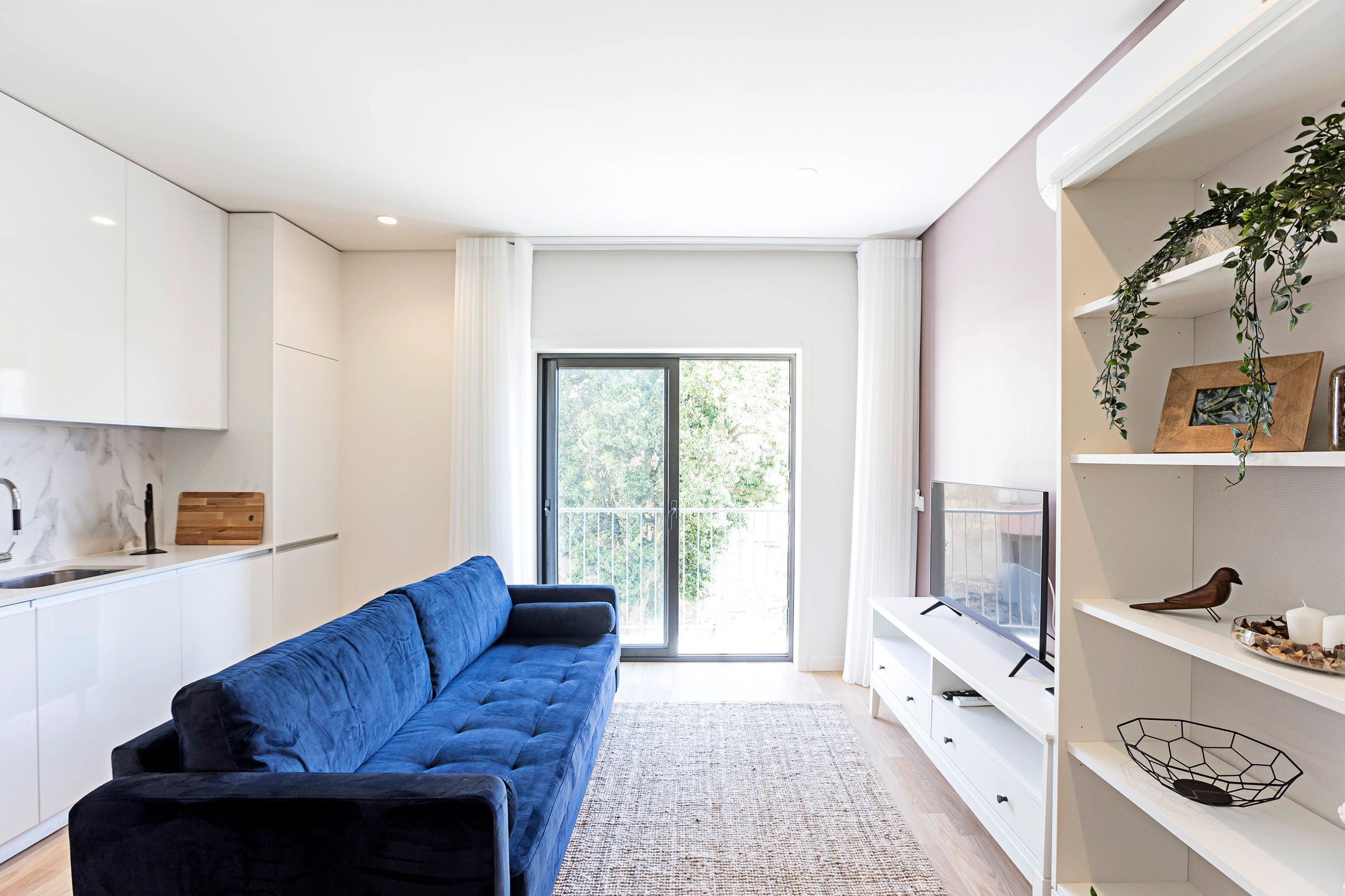 A modern living space with a blue sofa, white cabinetry, a large window, and a balcony.