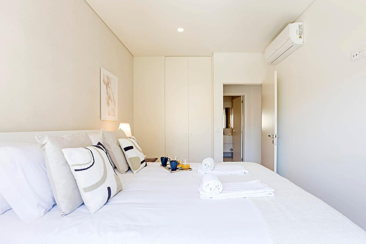 A hotel room with a bed, pillows, a bedside table with a lamp, a tray with breakfast items, and an air conditioning unit.