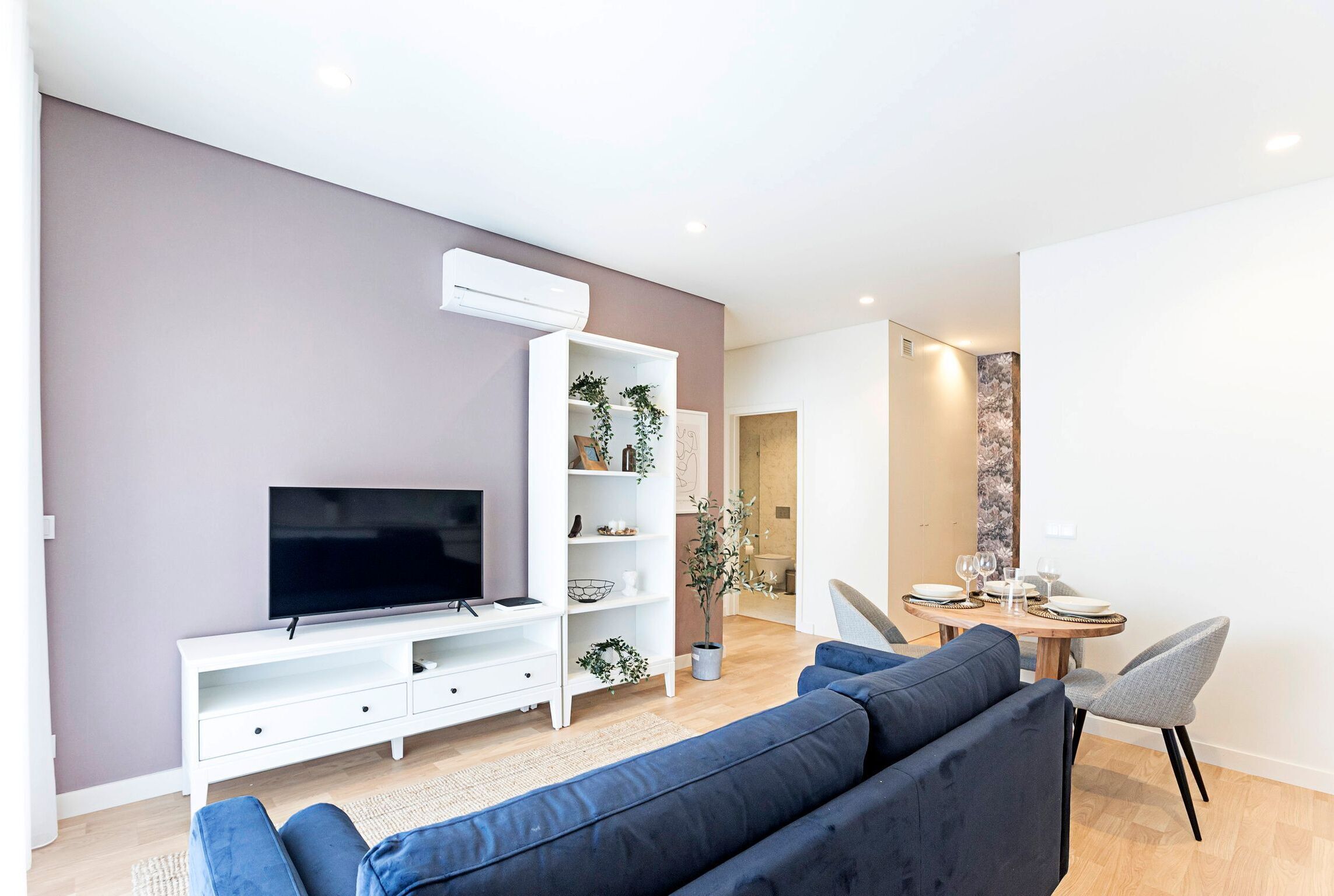 A modern living room with a flat-screen TV, a blue sofa, a white entertainment unit, and a dining area with a table and chairs.