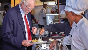 A man in a suit receiving food from a chef in a kitchen setting. - Hotel Zesper (Kisii)