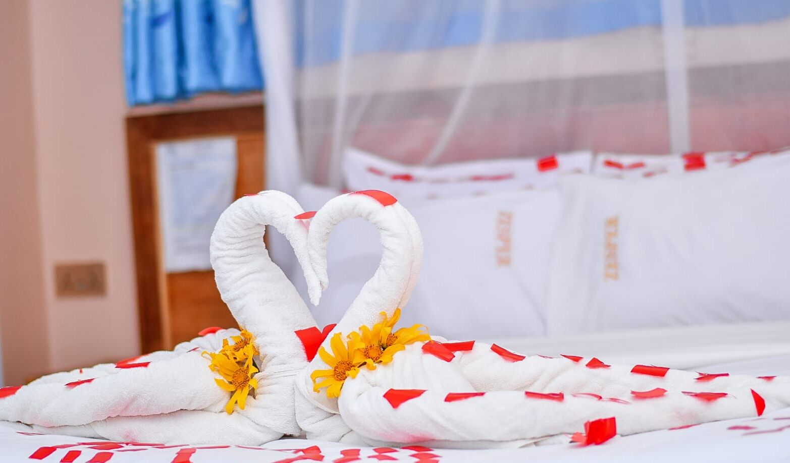 Un lit avec du linge de lit blanc et des touches de rouge, un cygne décoratif fait avec une serviette blanche, et un arrangement floral.