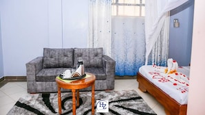 A room with a grey sofa, a small round wooden table with a tray of items, a bed with a white and red bedspread, and a window with a blue curtain. - Hotel Zesper (Kisii)