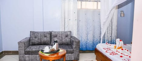 A room with a grey sofa, a small round wooden table with a tray of items, a bed with a white and red bedspread, and a window with a blue curtain.