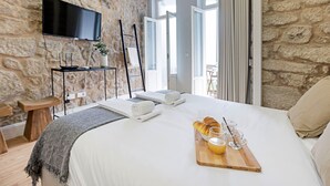 A hotel room with a stone wall, a flat-screen TV, a wooden side table, a bed with white linens, a tray with croissants and orange juice, and a view of the outdoors. - Cimo de Vila 62 by LovelyStay (Porto)