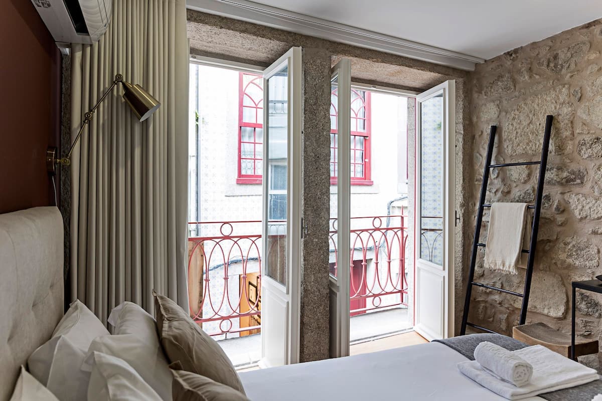 A hotel room with a bed, a bedside lamp, a window with a view of a balcony, and a stone wall.