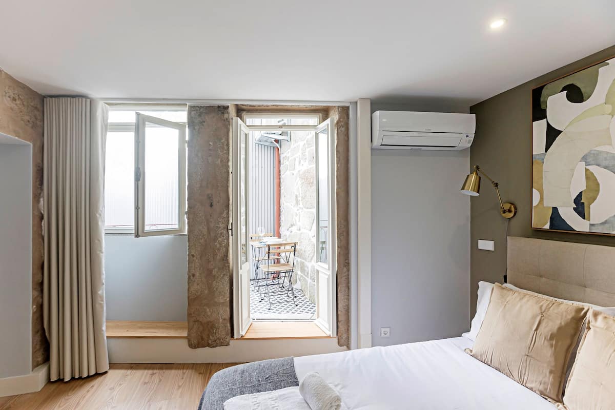 A bedroom with a bed, an air conditioner, a window with curtains, and a view of an outdoor area.