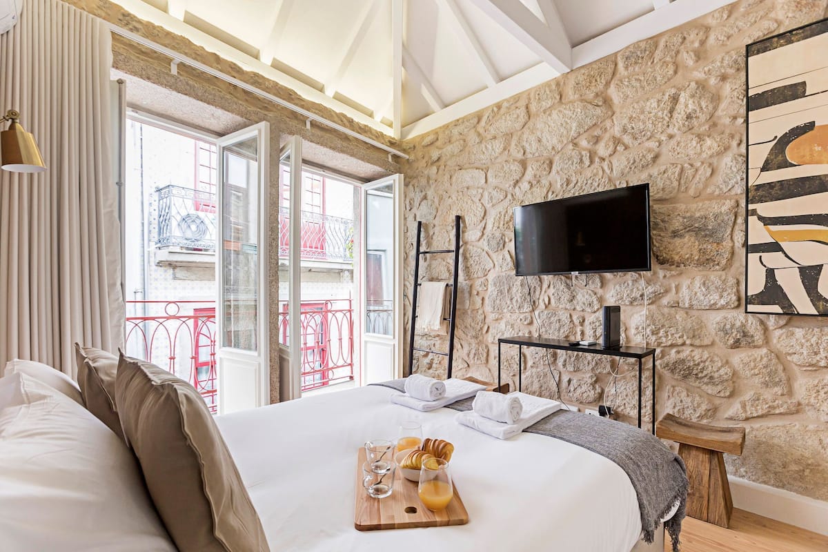A hotel room with a stone wall, a bed with a wooden headboard, a TV mounted on the wall, and a balcony with red railings.