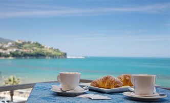 Una mesa con dos tazas de café y dos cruasanes sobre un mantel azul, con vistas a la costa y un pueblo lejano.