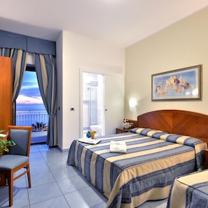 A hotel room with a bed, a desk, a chair, a balcony with a view, and a painting on the wall.