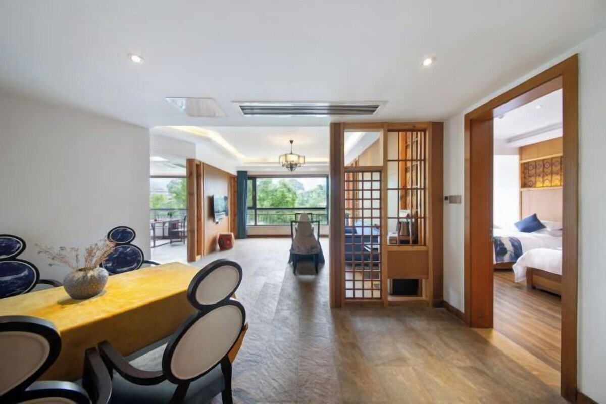 Une chambre d’hôtel moderne avec un plancher en bois, un coin repas avec une table et des chaises, une vue dégagée sur l’extérieur grâce à une grande fenêtre, et une porte coulissante de style japonais traditionnel.