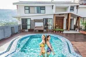 Une piscine où se trouvent deux personnes, une maison moderne avec un balcon et une vue sur les montagnes.