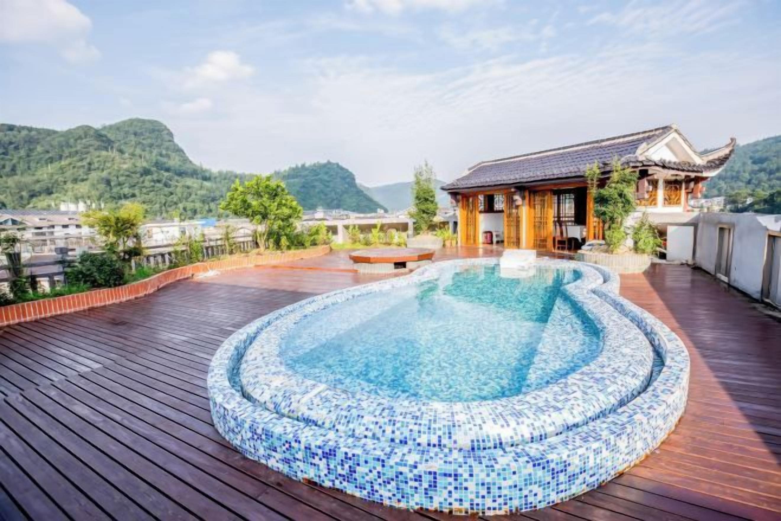 Une piscine sur le toit, bordée de carreaux, entourée d’une terrasse en bois, offrant une vue sur un bâtiment traditionnel et un paysage montagneux.