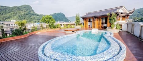 A rooftop pool with a tiled edge, surrounded by a wooden deck, overlooking a traditional building and a mountainous landscape.