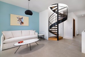A modern living room with a spiral staircase, a white sofa, a coffee table with fruit, and a painting on the wall.
