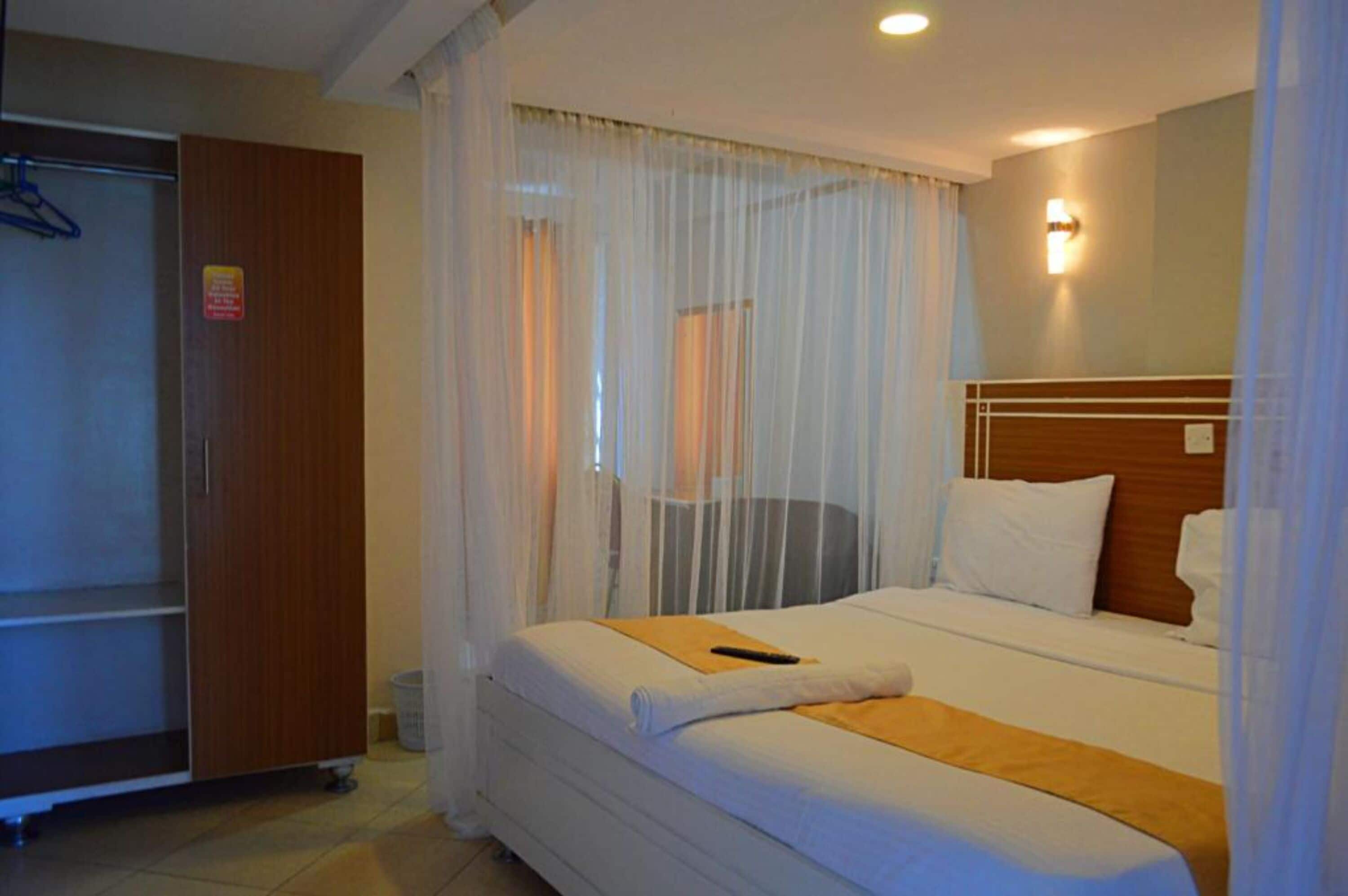 Une chambre d’hôtel comprenant un lit, une tête de lit en bois, une table de chevet, une commode, une armoire et une fenêtre avec des rideaux légers.