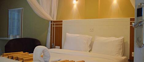A hotel room with a bed, white linens, a headboard, two wall sconces, a window with curtains, and a bedside table with a lamp.