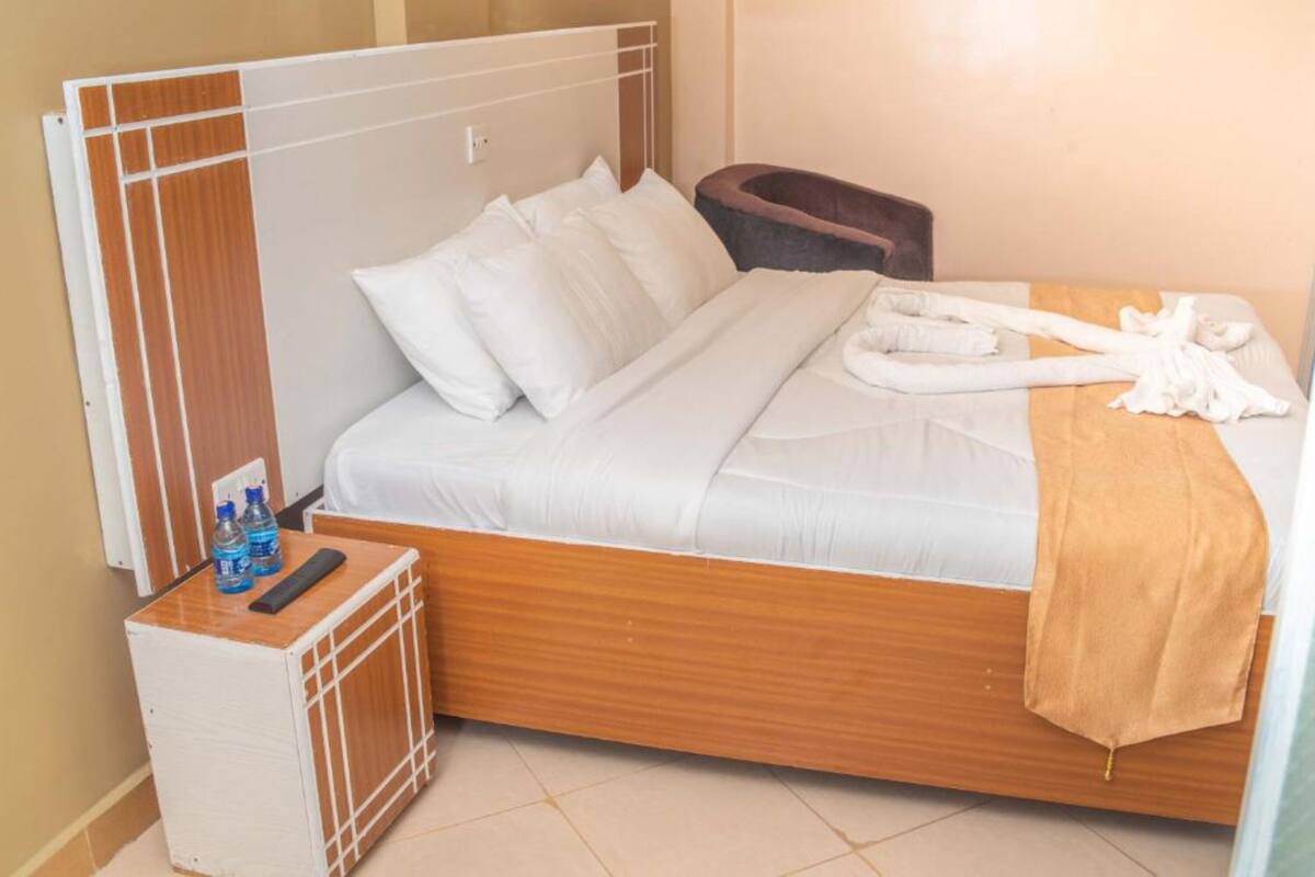 A hotel room with a wooden bed, white bedding, a small bedside table with bottles, and a brown bath mat.