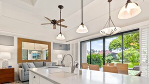 A modern kitchen with a large island, white cabinetry, and a view of a lush garden through large windows. - Tranquil Retreat w/Split AC - Kiahuna 2052 (KOLOA)