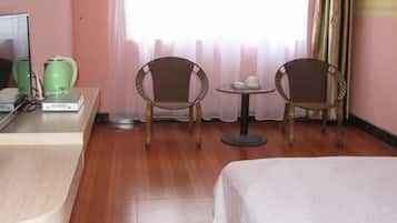 Habitación de hotel con una cama, dos sillones de mimbre, una mesita redonda con tetera y tazas, un televisor y una tetera verde sobre un escritorio.