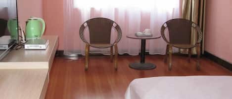 Habitación de hotel con una cama, dos sillones de mimbre, una mesita redonda con tetera y tazas, un televisor y una tetera verde sobre un escritorio.