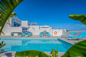 A swimming pool with blue lounge chairs and umbrellas, surrounded by white buildings and greenery.