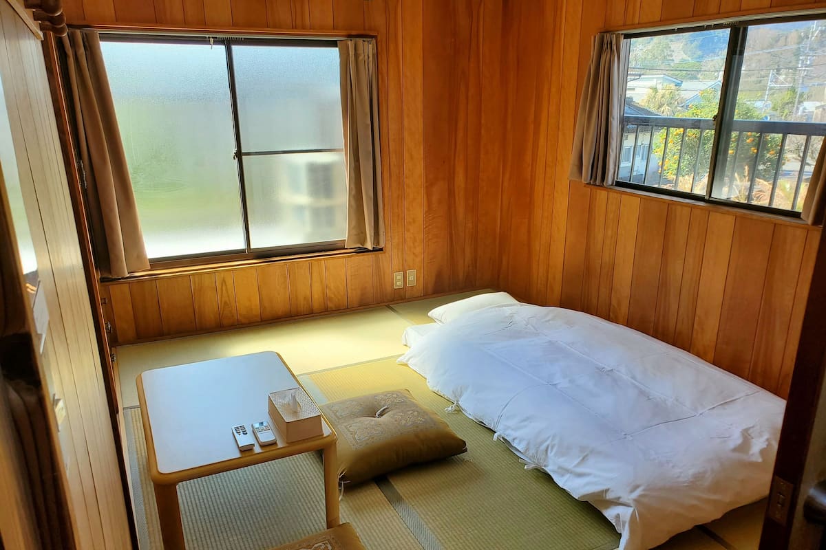 A small, cozy room with a bed, a table, and a chair. There are two windows with curtains, and the room has wooden walls.