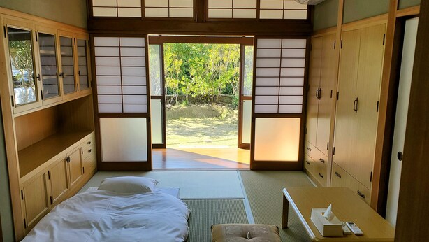 A traditional Japanese-style room with tatami flooring, a bed with white bedding, a small table with a tissue box, and sliding doors leading to a garden. - SEA-DWELLER Inn (Miyazaki)