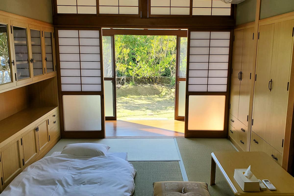 A traditional Japanese-style room with tatami flooring, a bed with white bedding, a small table with a tissue box, and sliding doors leading to a garden.