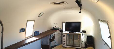 A compact RV interior with a television, a small kitchenette, a fur rug, and a wooden shelf.