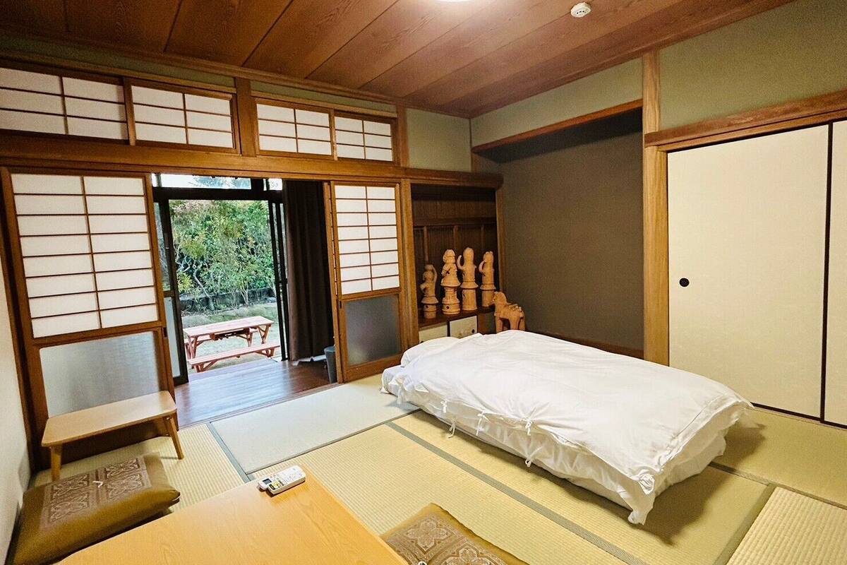 A traditional Japanese room with tatami flooring, a bed, a wooden table, and a sliding door leading to an outdoor area.