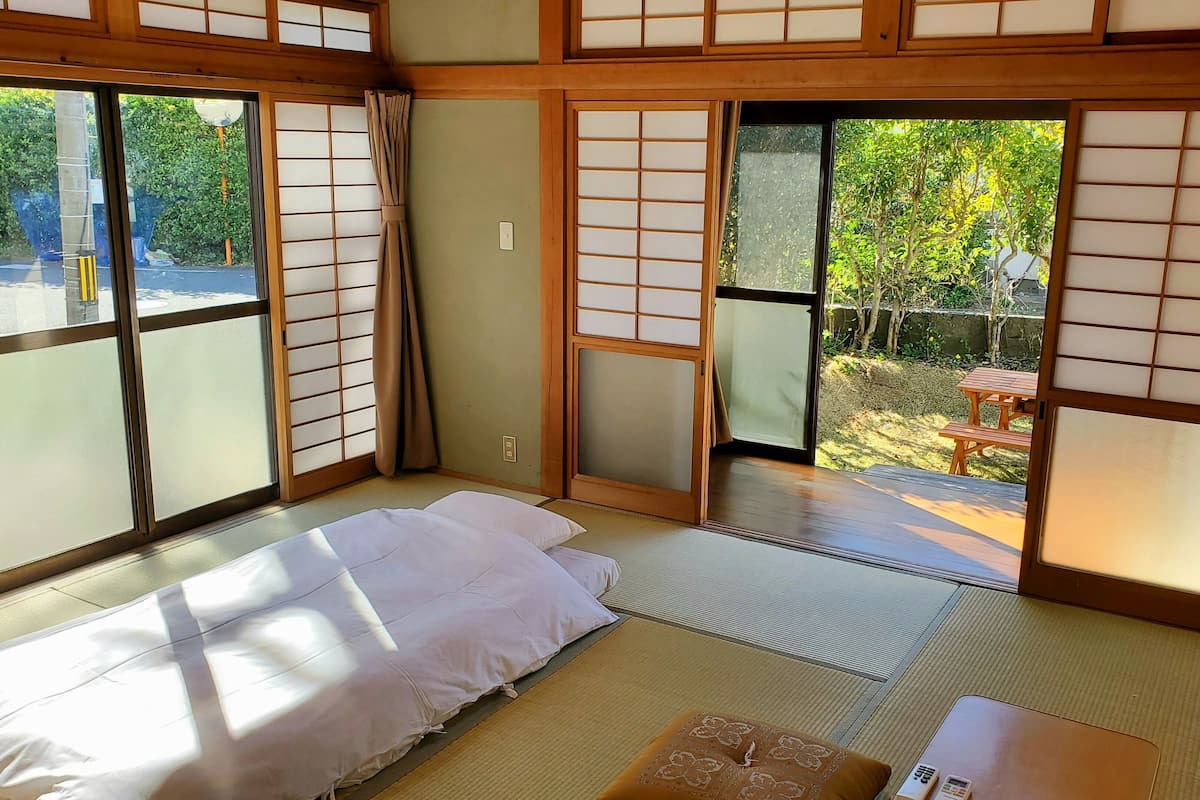A traditional Japanese room with tatami flooring, a bed with white bedding, sliding shoji doors, and a view of a garden.