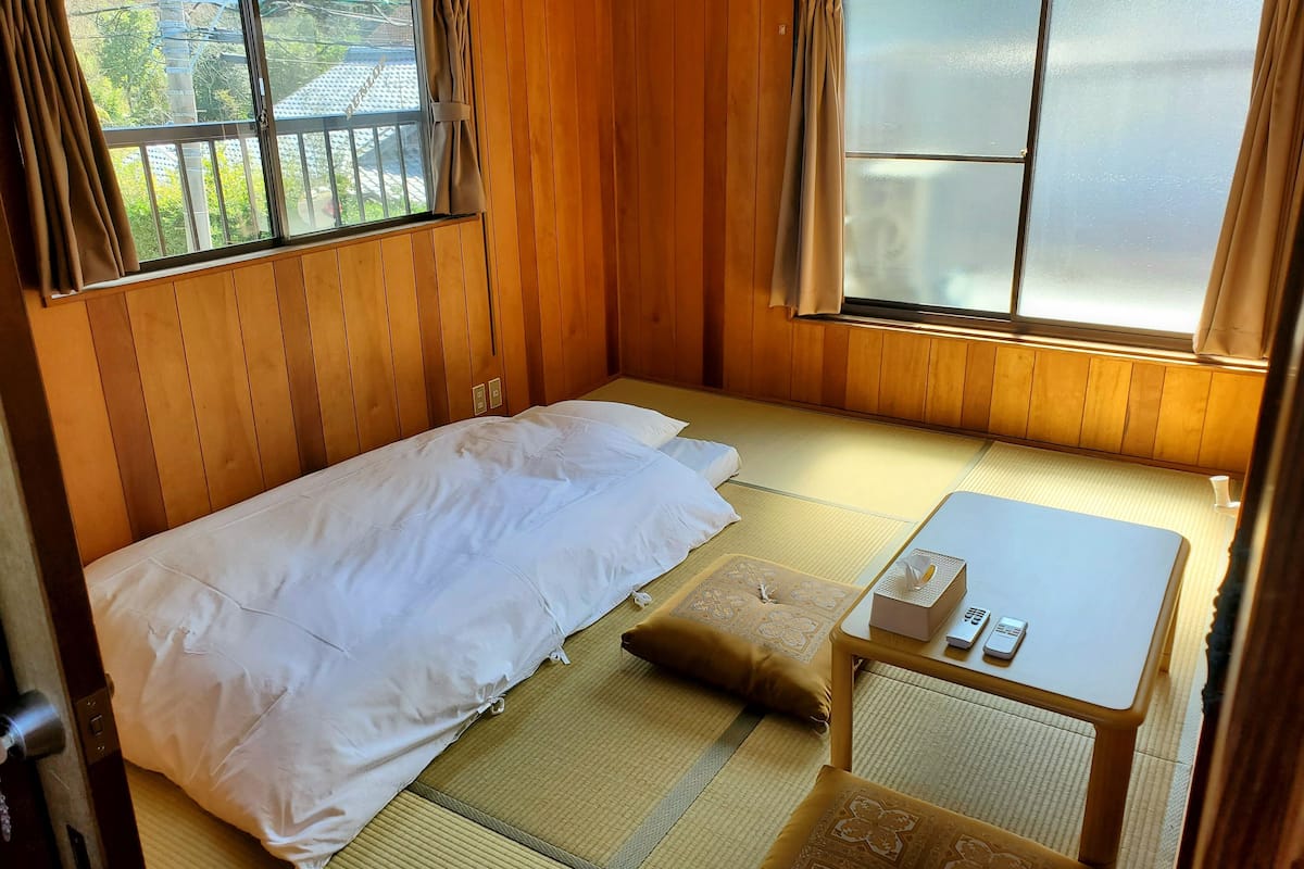 A small, cozy room with a bed, a small table, and two windows.
