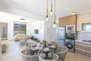 A modern kitchen with a built-in oven, microwave, and washing machine, a dining area with a round table and chairs, and a living room with a sofa and coffee table.