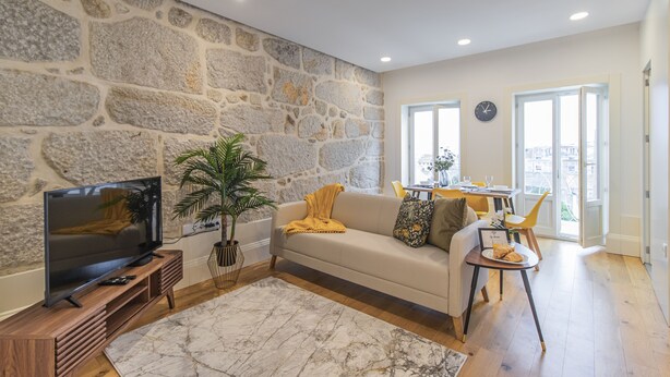 Un salon avec un mur en pierre, un téléviseur posé sur un meuble en bois, un canapé avec des coussins, une table basse et un coin repas avec des chaises et une table.