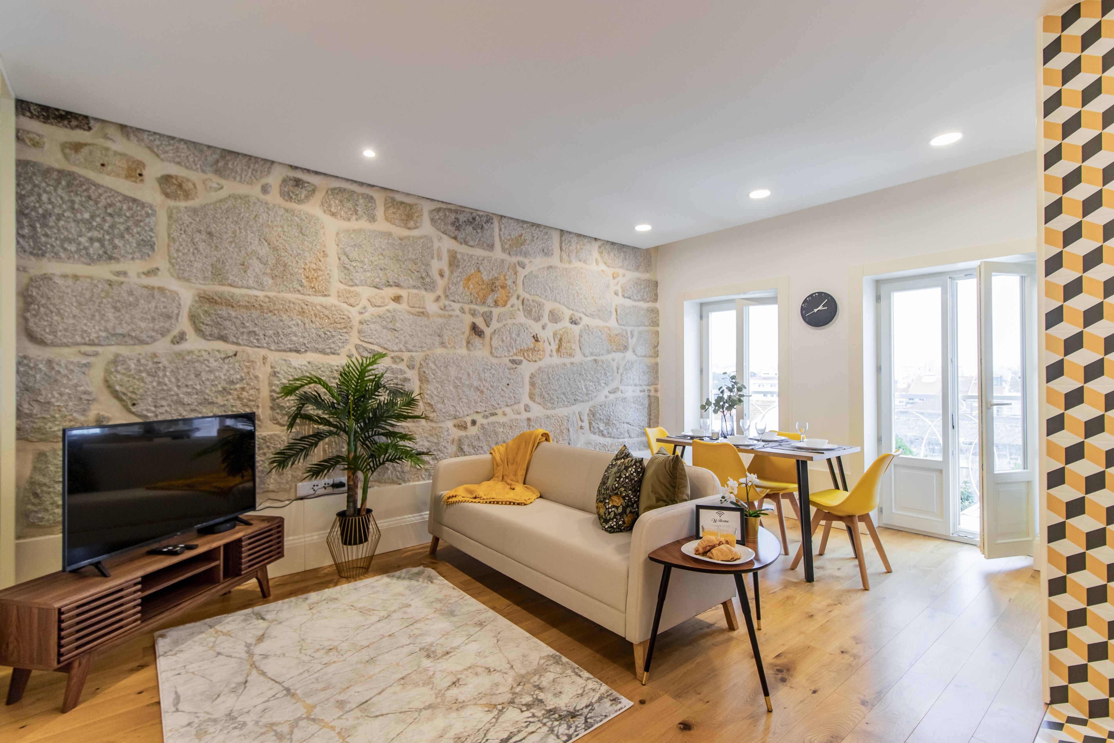 Un salon avec un mur en pierre, un canapé, un téléviseur, un coin repas avec des chaises et une vue à travers une porte vitrée.