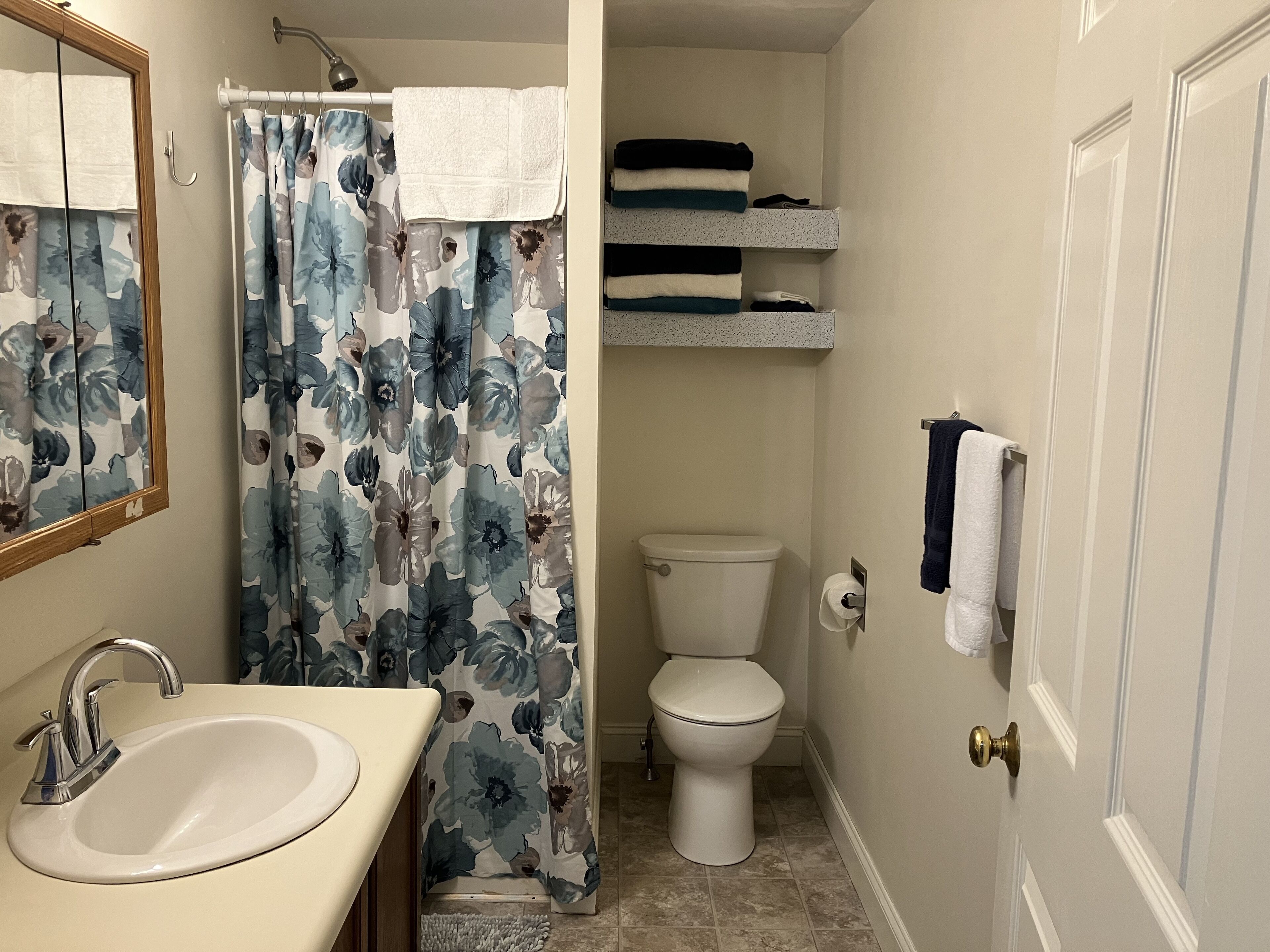 A bathroom with a sink, toilet, shower curtain, and shelves with towels.