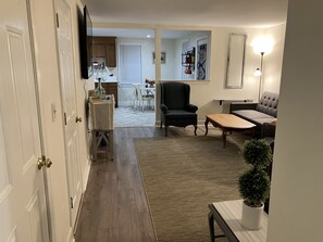 Een woonkamer met een bank, fauteuil, salontafel en een kamerplant. Achtergrond toont een keuken met kasten en een eettafel.